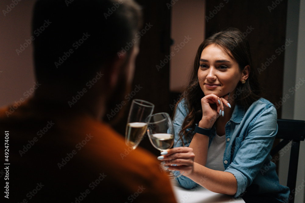 Close-up back view to smiling pretty young woman talking celebration toast and clinking white wine glass with husband during romantic dinner. Love couple celebrating anniversary or Valentines day.