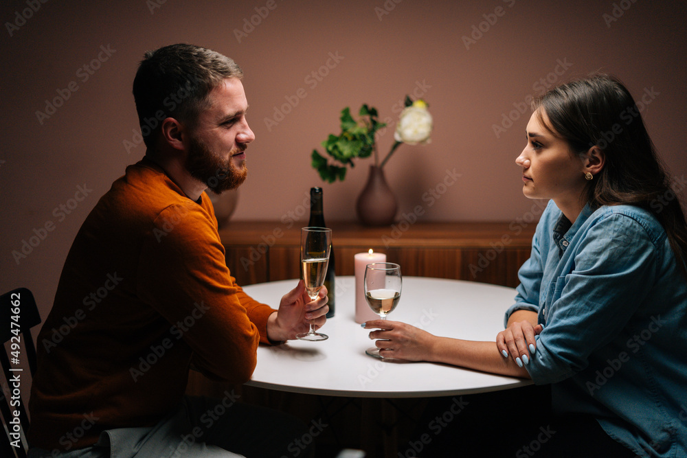 Side view of happy romantic young couple enjoying talking and sitting together at dinner table with candles in dark room. Love man and woman celebrating anniversary, Valentines day at home. Side view of happy romantic young couple enjoying talking and sitting together at dinner table with candles in dark room. Love man and woman celebrating anniversary, Valentines day at home.