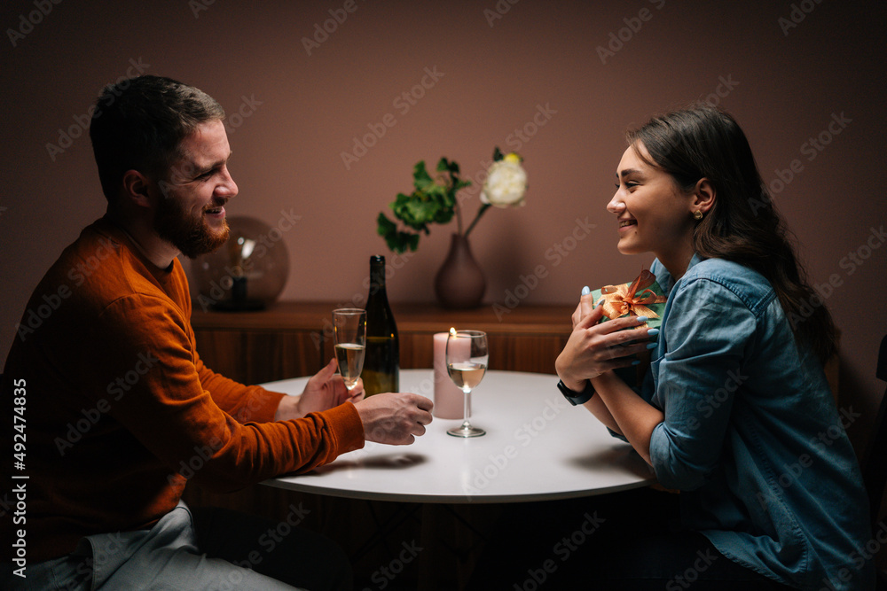 Loving boyfriend giving box with gift to charming happy girlfriend sitting at table with candles on birthday or Valentines Day. Happy wife receiving present from husband enjoying romantic dinner date.