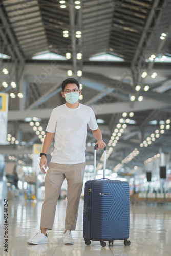 Young male wearing face mask with luggage walking in airport, protection Coronavirus disease infection, Asian man traveler with hat. Time to travel after vaccine booster dose concept