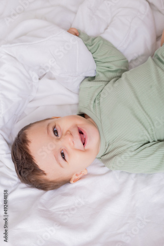 portrait of a cute smiling baby in a green cotton bodysuit lying on a white bed on his back