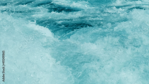 Close-up, strong splash of sea turquoise water from fast sailing motor boat. Powerful white foam waves rise, break into splashes. Huge stream of crystal clear green tide water in ocean