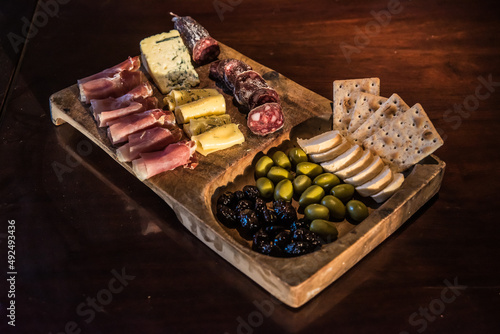 charcuterie board, with cheeses, crackers, salami, prosciutto, roquefort cheese, green and black olives, on a wooden board, on a polished wooden table.