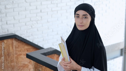 A woman student in a hijab holds books in her hands and looks into the frame. The concept of education for Muslim women who wears a hijab. Beautiful muslim woman in black hijab at university