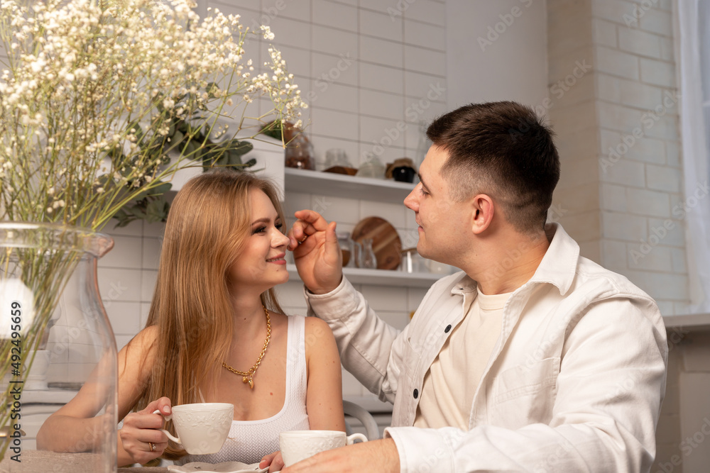 Young couple sitting at kitchen table, holding cups with tea or coffee, looking at each other and talking. Happy wife and husband. High quality photo