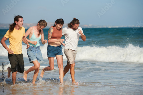 Cheerful young men and women running on beach and splashing water on sunny day