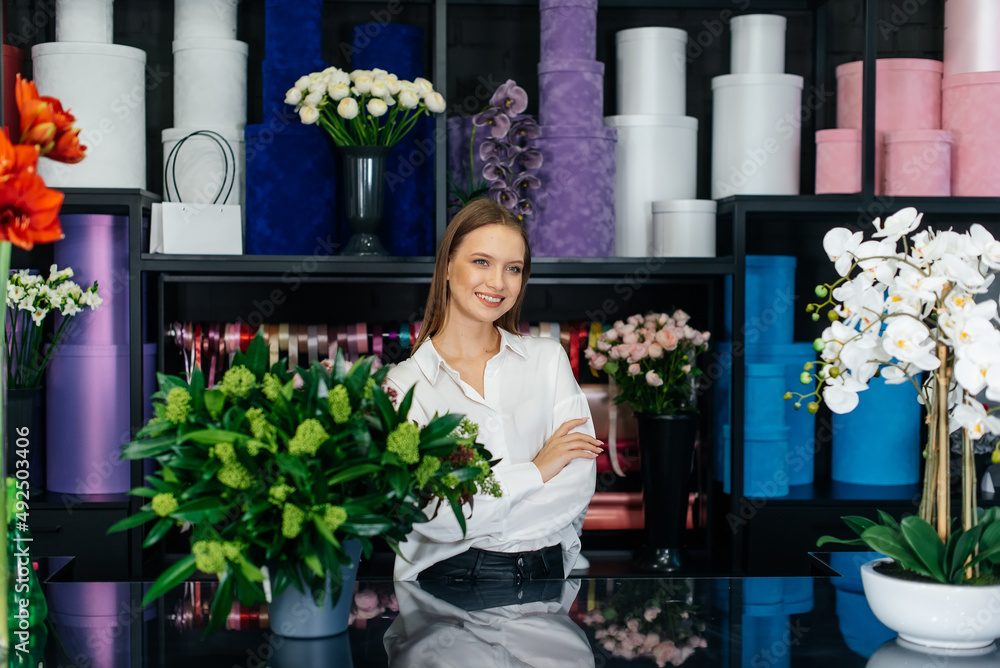 Fototapeta premium A young female florist poses and smiles against a background of flowers in a cozy flower shop. Floristry and making of bukets in a flower shop. Small business