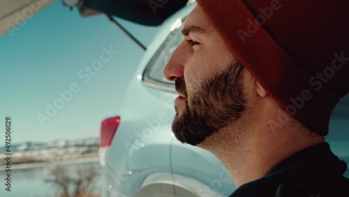 A close up of a millennial male overlooks a gorgeous winter lake underneath a camping awning.  The suv reflects a stunning blue sky and sun flares.  The relaxing male takes a swig of bottled beer.