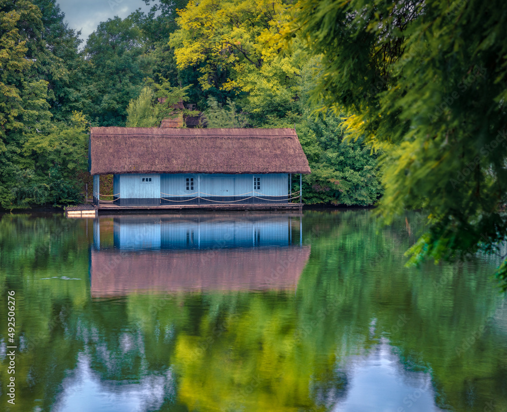 Small fishing chalet with straw roof among green fresh lievs in King Mihai I of Romania Park (Herastrau Park), Bucharest, Romania, Europe. Beauty of countryside concept background. Small fishing chalet with straw roof among green fresh lievs in King Mihai I of Romania Park (Herastrau Park), Bucharest, Romania, Europe. Beauty of countryside concept background.
