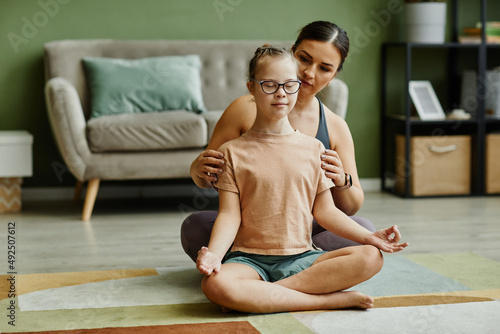 Portrait of female instructor assisting young girl with down syndrome doing yoga and meditating in fitness studio, copy space