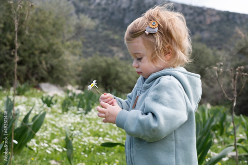 Funny cute adorable caucasian blonde baby girl, toddler in green field of daisy, camomiles smelling flowers.Curious child,kid, infant,learning environment,nature,mountains in spring or summer
