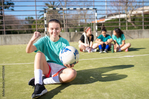 Portrait of happy teenage football girl with ball on stadium. Smiling girl sitting on field and other girls behind her, showing her fist, believing in victory. Healthy lifestyle and team sport concept