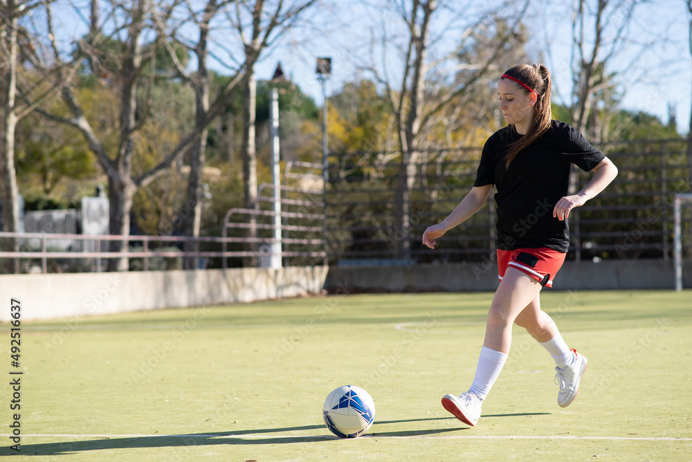 Beautiful girl joyfully playing football. Young girl in sport uniform training walking on field on sunny day, looking at ball and preparing to kick. Sport activity, active rest and hobby concept