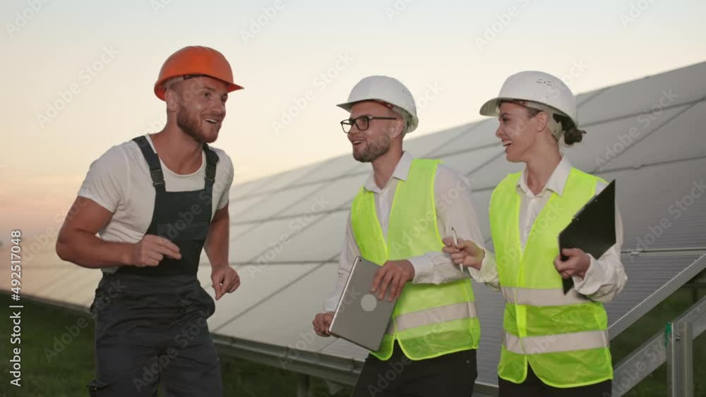 Professional technician and two inspectors in helmets and uniform dancing and smiling on station with solar panels. Three colleagues having fun during evening time after productive work. Professional technician and two inspectors in helmets and uniform dancing and smiling on station with solar panels. Three colleagues having fun during evening time after productive work.