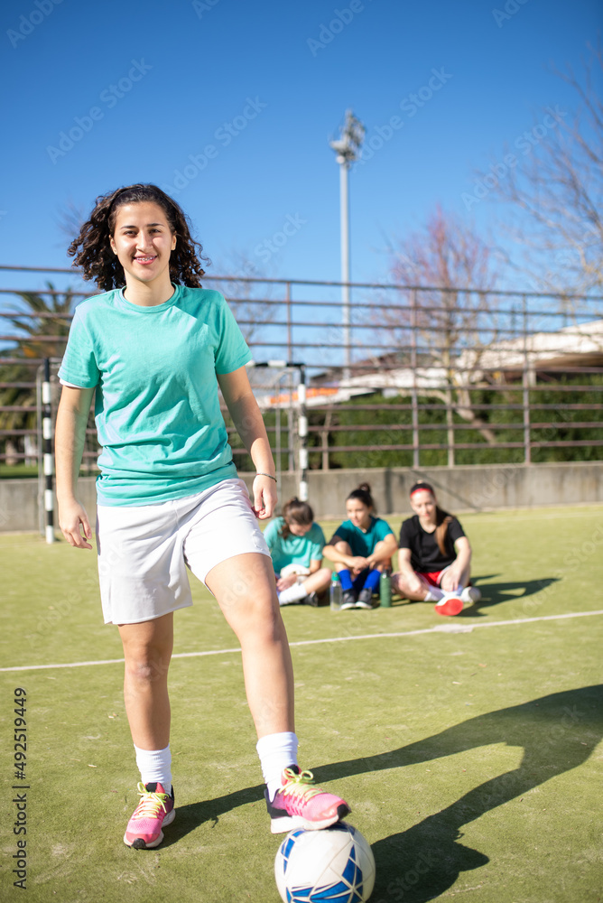Portrait of football girl with ball after game. Smiling girl standing on field with ball under her leg, posing for camera when other girls behind enjoying rest. Leisure activity and team sport concept