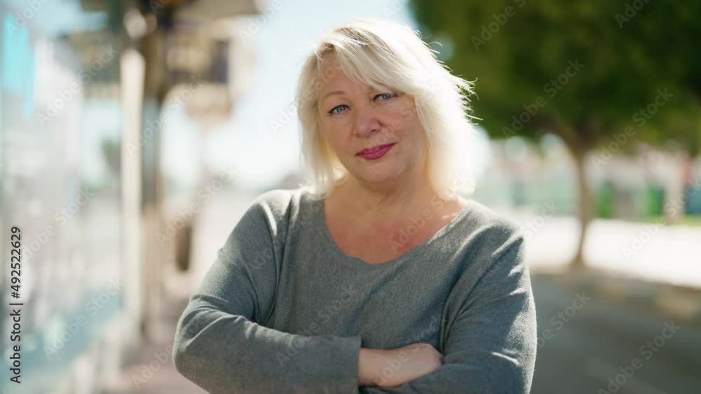 Middle age blonde woman with relaxed expression standing with arms crossed gesture at street Middle age blonde woman with relaxed expression standing with arms crossed gesture at street