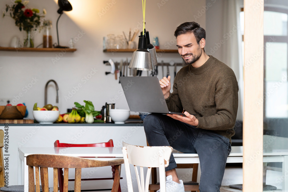 Smiling young man using laptop siting on table in kitchen at home Smiling young man using laptop siting on table in kitchen at home
