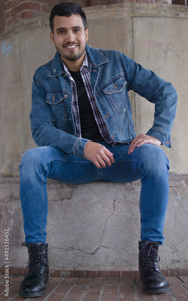 full length image of a Colombian man dressed in blue sitting on a concrete seat and smiling while looking at the camera full length image of a Colombian man dressed in blue sitting on a concrete seat and smiling while looking at the camera