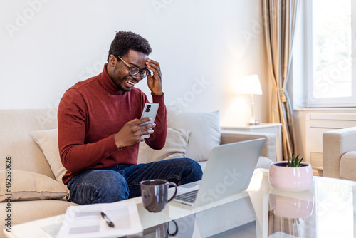 Excited Black African American Man Having a Video Call on Smartphone while Sitting on a Sofa in Living Room. Happy Man Smiling at Home and Talking to His Friends and Family Over the Internet.....