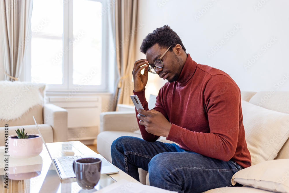 African man sit on couch using smartphone shocked by reading online news, surprised male get breaking message or text on mobile phone, confused guy holding cell see warning notification