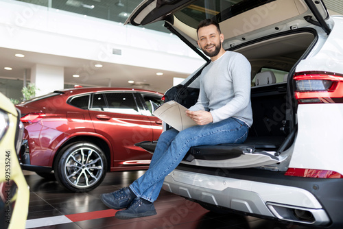 a buyer studying a booklet in a showroom of a car dealership sitting in the trunk of a crossover looking at the camera with a smile