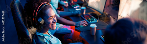 Cheerful African female cybersport gamer wearing wired headset sitting on gaming chair and watching game of teammate in cyber club photo