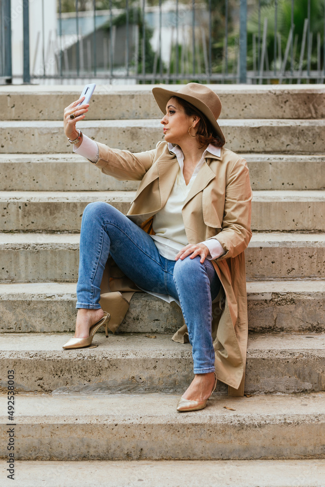 An elegant adult woman sitting on steps outdoors and taking a selfie