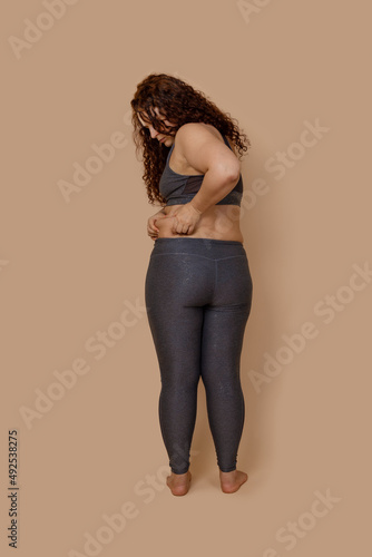 Vertical photo of rear barefoot concerned melancholic fat obese woman, looking on dangling hips, shove in leggings. Wearing gray sportswear. Sad looking, big size. Body positive, loosing motivation