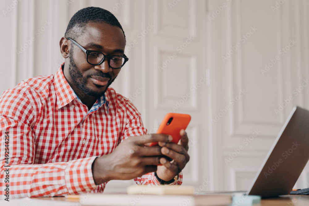 Young handsome african male executive in glasses sitting at office table and using mobile phone Young handsome african male executive in glasses sitting at office table and using mobile phone
