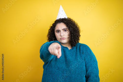 Young beautiful woman wearing a birthday hat over isolated yellow background pointing with finger to the camera and to you, confident gesture looking serious © Irene