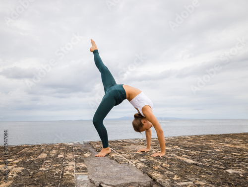 Slim Caucasian woman practicing gymnastic exercise. Wheel pose one leg up. Fitness, wellness concept. Flexible body. Outdoor activities. Cloudy sky background. Copy space. Bali