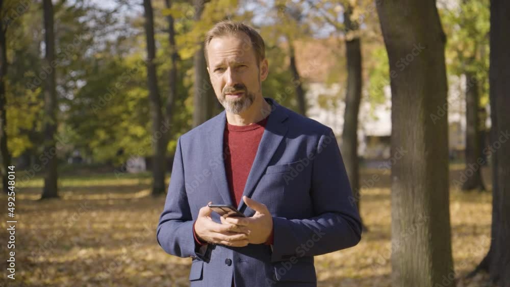 A middle-aged handsome Caucasian man talks to the camera and holds a smartphone in a park in fall - trees in the blurry background A middle-aged handsome Caucasian man talks to the camera and holds a smartphone in a park in fall - trees in the blurry background