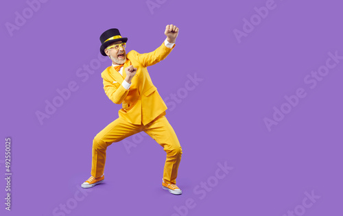 Funny senior man fighting imaginary enemy. Full body shot of happy crazy silly old man wearing yellow suit and top hat fighting invisible opponent and having fun isolated on solid purple background