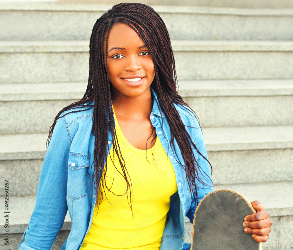 Fototapeta premium Portrait of happy smiling young woman with skateboard wearing vivid yellow hat in the city