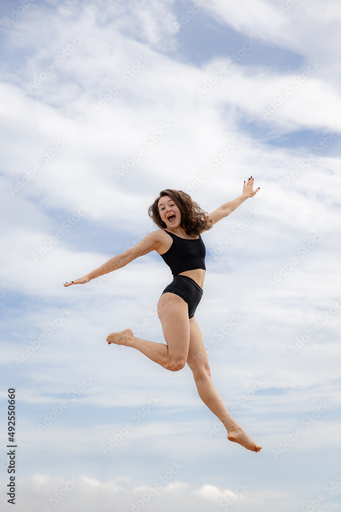 Excited young woman jumping over cloudy blue sky. Caucasian woman wearing black sportswear. Fitness, wellness concept. Outdoor activity. Copy space. Sky background. Bali