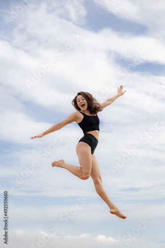 Excited young woman jumping over cloudy blue sky. Caucasian woman wearing black sportswear. Fitness, wellness concept. Outdoor activity. Copy space. Sky background. Bali