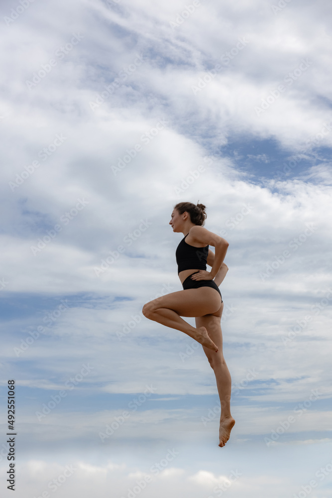 Slim sportive woman jumping over cloudy blue sky. Young Caucasian woman wearing black sportswear. Fitness, wellness concept. Outdoor activity. Copy space. Sky background. Bali