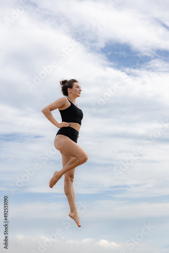 Fototapeta Naklejka Na Ścianę i Meble -  Slim gymnast woman jumping over cloudy blue sky. Young Caucasian woman wearing black sportswear. Fitness, wellness concept. Outdoor activity. Copy space. Sky background. Bali