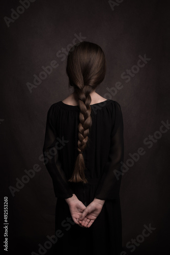 Rear view of girl with long hair in braid and hands on back in classic dark studio portrait photo
