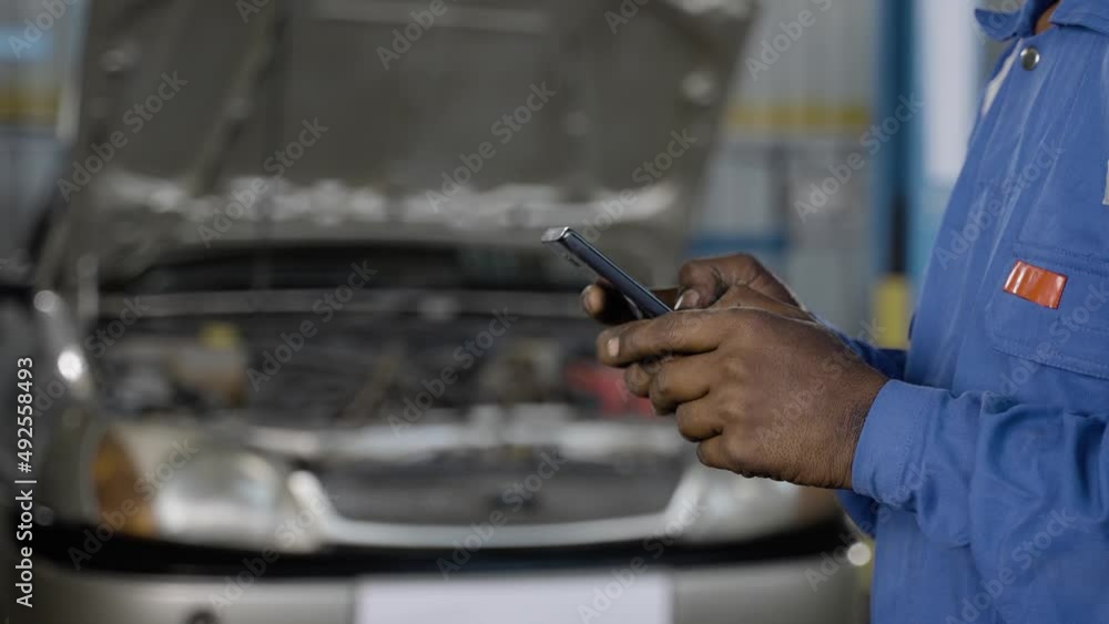 Close up of mechanic hands busy using mobile phone in front of broken car at garage or workshop - concept of communication, internet and technology Close up of mechanic hands busy using mobile phone in front of broken car at garage or workshop - concept of communication, internet and technology
