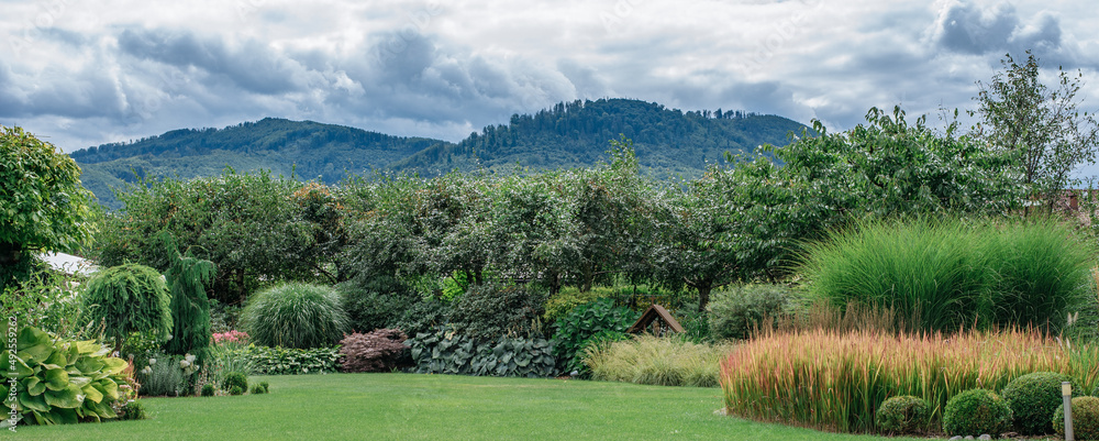 Naklejka premium Piękny zadbany ogród na tle gór w malowniczym miejscu. Stylowy ogród pełen kwiatów i zieleni oraz traw ozdobnych
