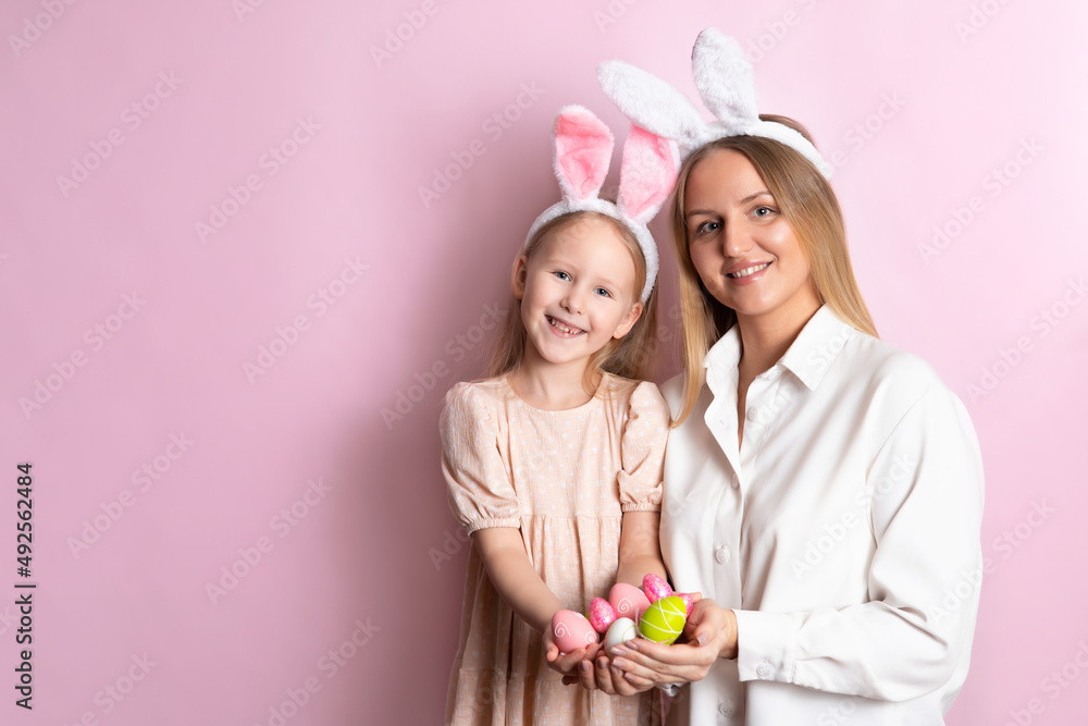 Happy Easter. Portrait of beautiful blonde mother and daughter with rabbit ears and a basket of colored eggs in their hands. Pink background. Space for text. Happy Easter. Portrait of beautiful blonde mother and daughter with rabbit ears and a basket of colored eggs in their hands. Pink background. Space for text.