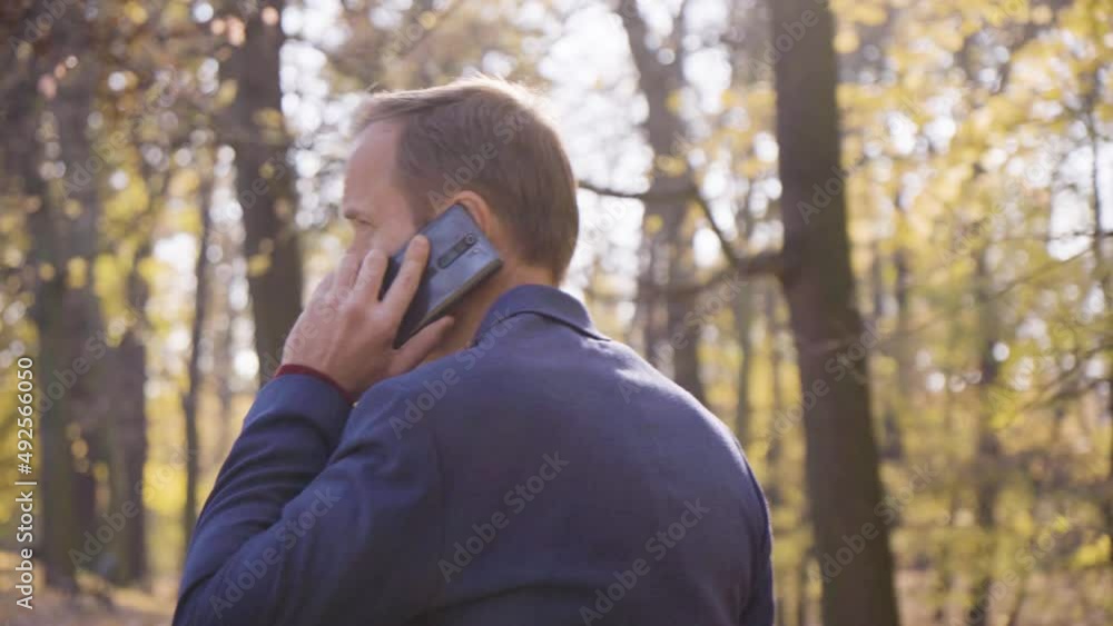 A middle-aged handsome Caucasian man talks on a smartphone as he walks in a park in fall - rear closeup - tree crowns in the blurry background A middle-aged handsome Caucasian man talks on a smartphone as he walks in a park in fall - rear closeup - tree crowns in the blurry background