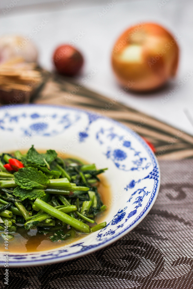 Stir Fried Morning Glory with Soybean Paste Served in a Chinese-patterned dish with spotted leaves, onions, shallots on the table.