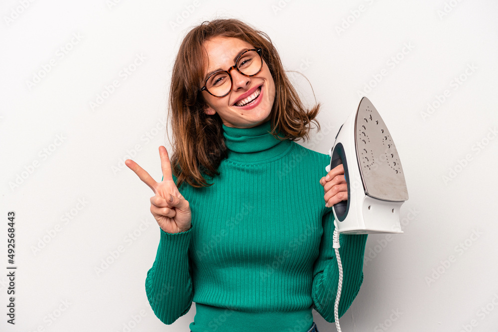 Young caucasian woman holding iron isolated on white background joyful and carefree showing a peace symbol with fingers. Young caucasian woman holding iron isolated on white background joyful and carefree showing a peace symbol with fingers.