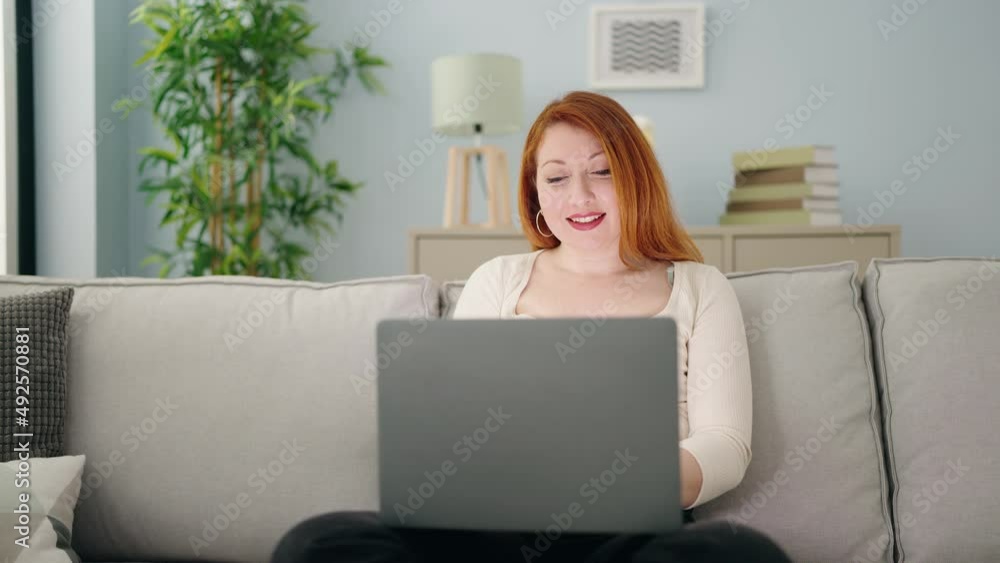 Young redhead woman using laptop sitting on sofa at home Young redhead woman using laptop sitting on sofa at home