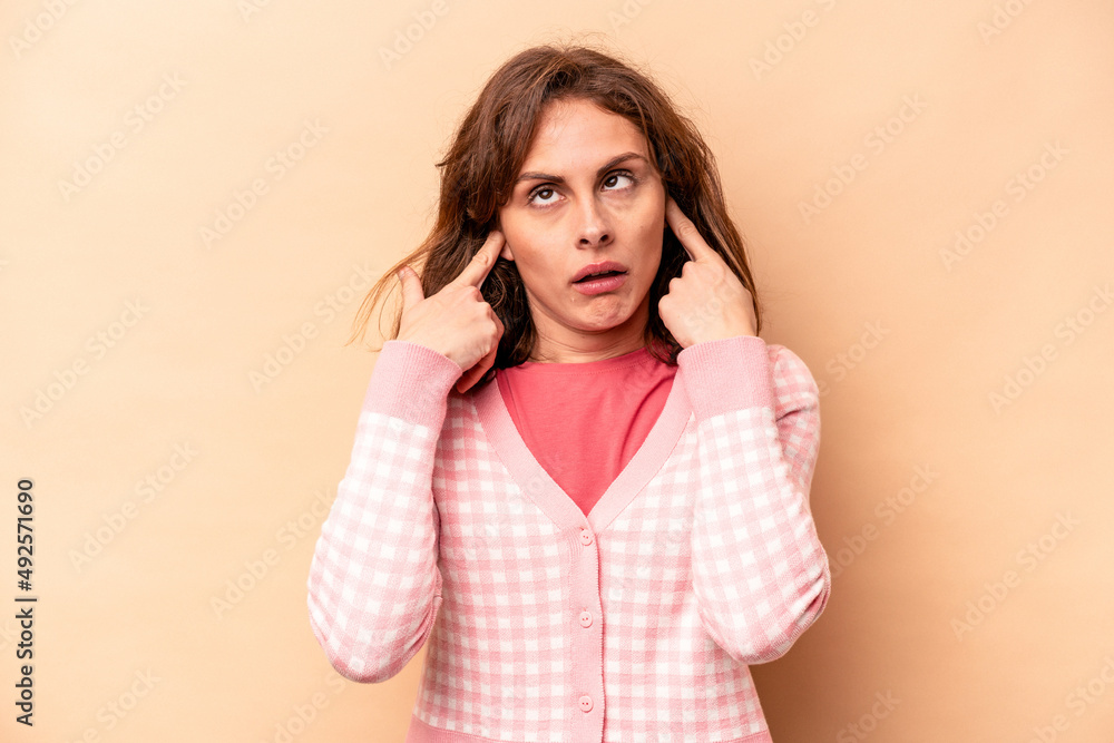 Young caucasian woman isolated on beige background covering ears with fingers, stressed and desperate by a loudly ambient. Young caucasian woman isolated on beige background covering ears with fingers, stressed and desperate by a loudly ambient.