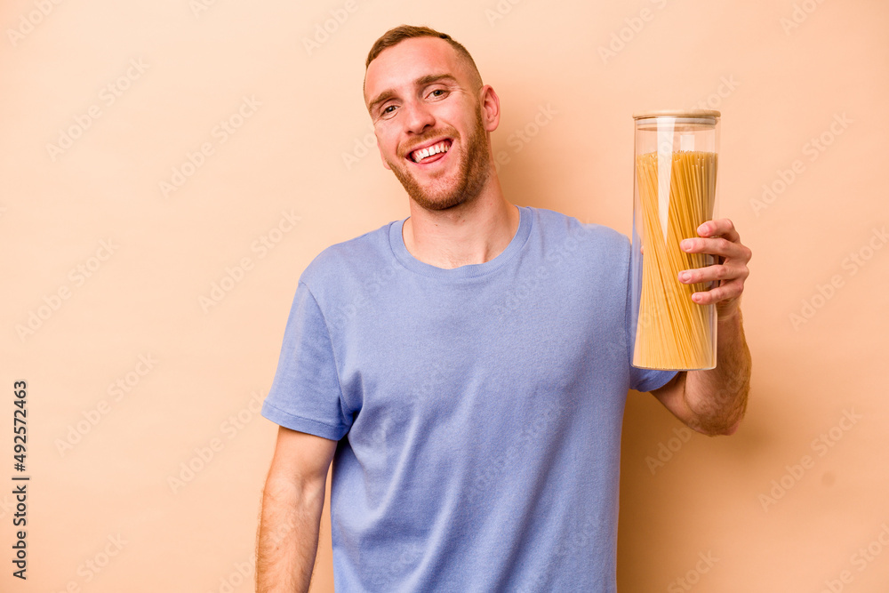 Young caucasian man holding spaghettis jar isolated on beige background laughs out loudly keeping hand on chest.