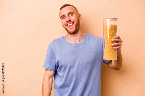 Young caucasian man holding spaghettis jar isolated on beige background laughs out loudly keeping hand on chest.