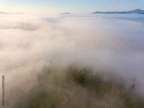 Fog envelops the mountain forest. The rays of the rising sun break through the fog. Aerial drone view.